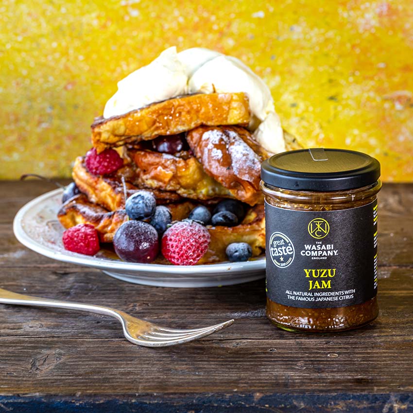 A plate of French toast topped with whipped cream and mixed berries is displayed beside a jar of yuzu jam on a wooden table. A fork rests in the foreground.