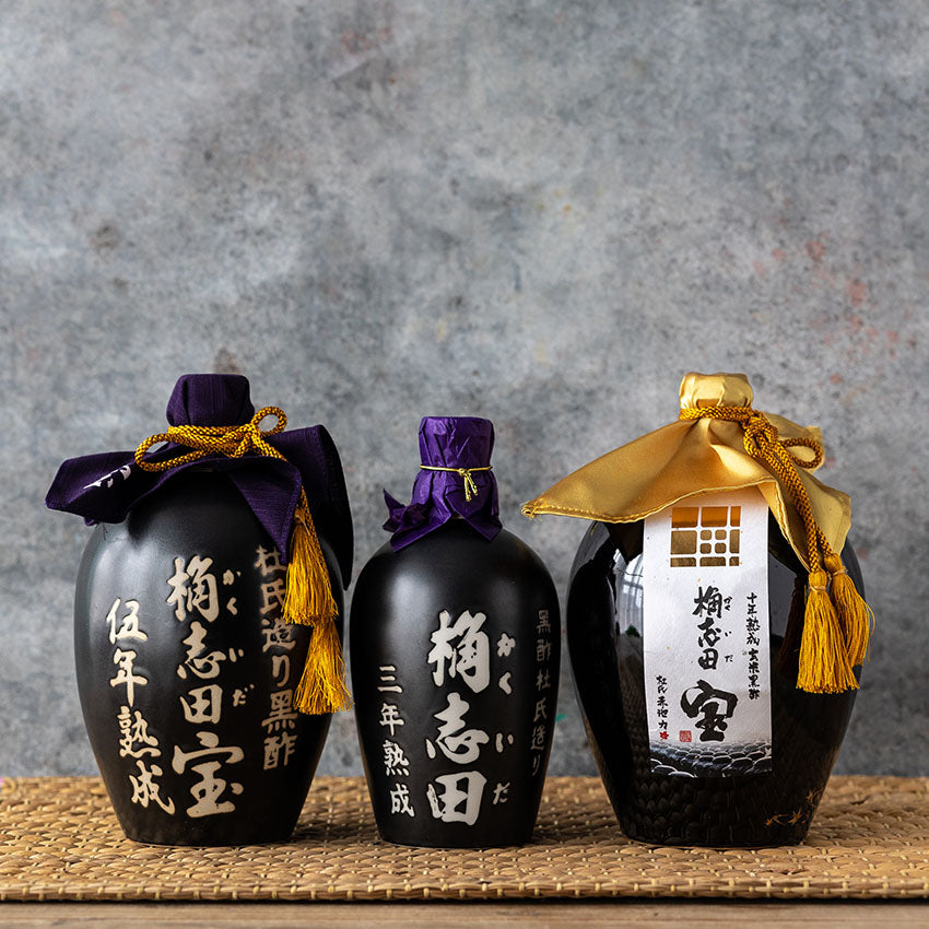 Three black ceramic sake bottles with Japanese calligraphy, each topped with colored cloth and decorative cord, are displayed on a wooden surface against a gray textured background.