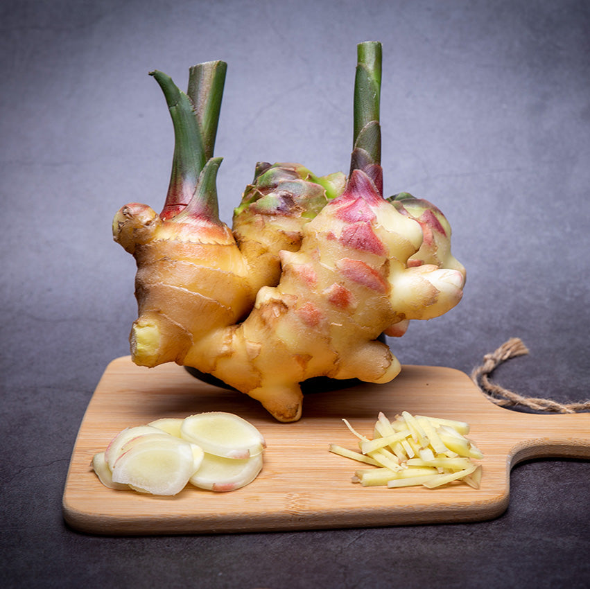 A whole ginger root with green stems sits on a wooden cutting board, alongside sliced and julienned pieces of ginger.