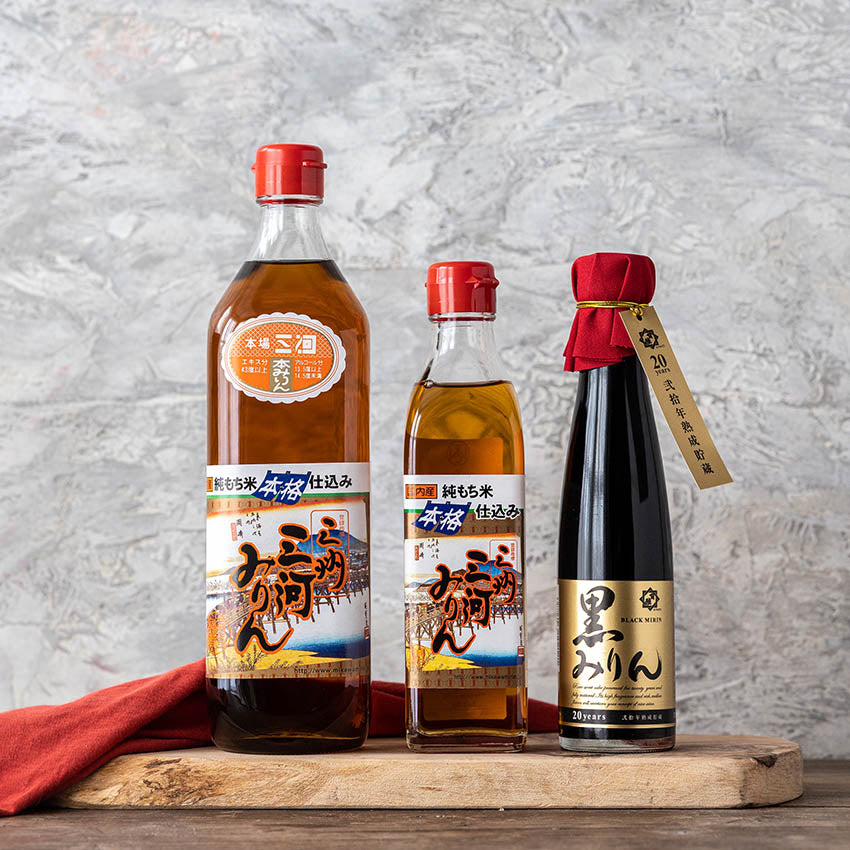 Three bottles of Japanese soy sauce with different shapes and labels are arranged side by side on a wooden surface in front of a light textured background.