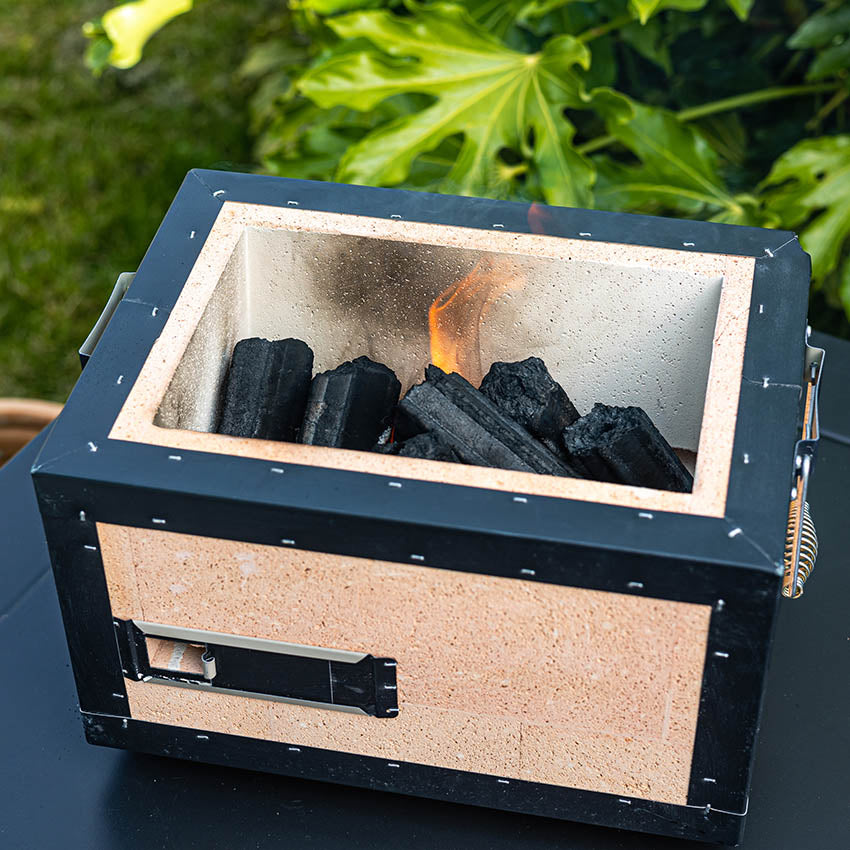 A small rectangular charcoal grill with burning charcoal inside, placed outdoors on a black table with green plants in the background.