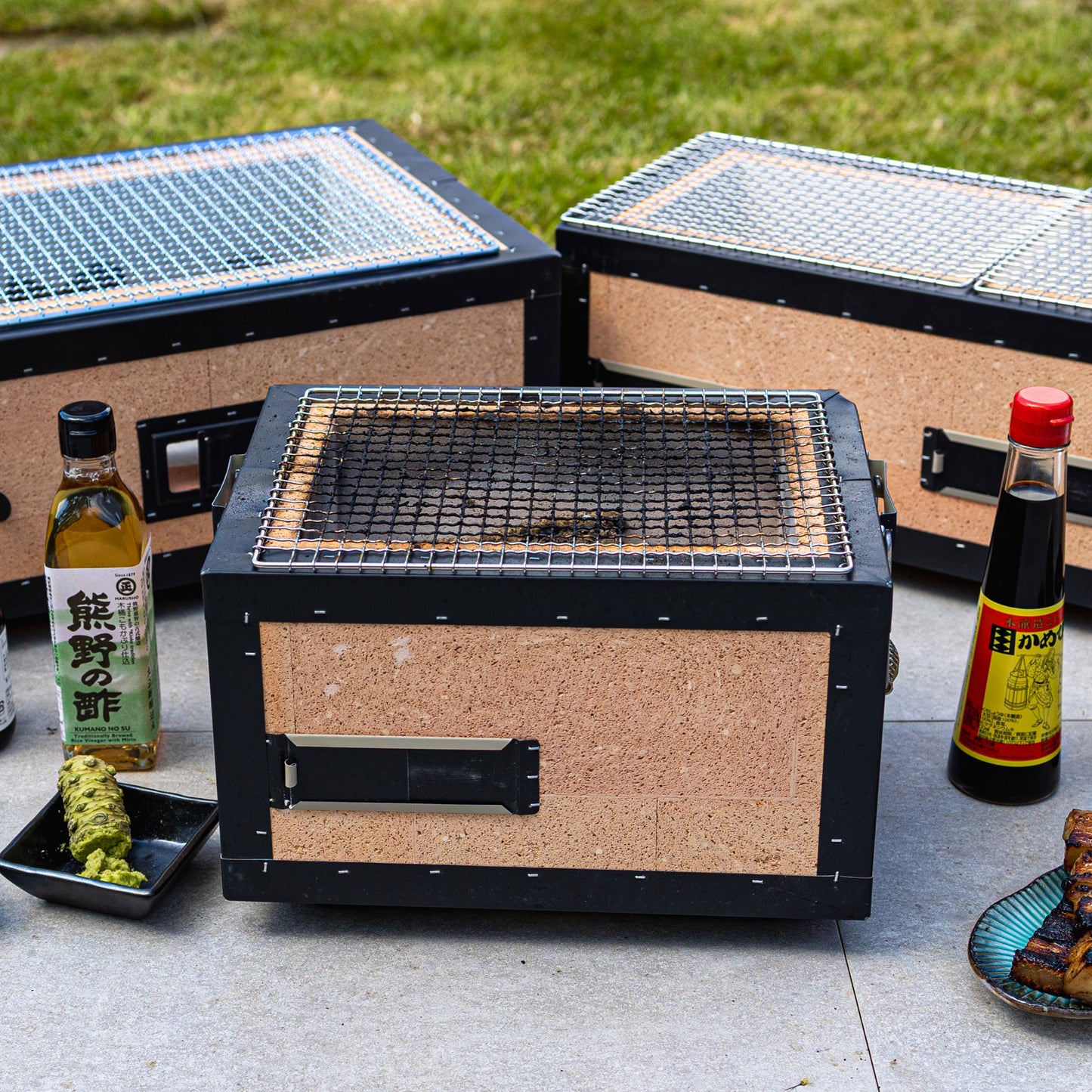 A rectangular tabletop grill sits outdoors, surrounded by bottles of Japanese condiments, a small dish of wasabi, and plates of grilled food.