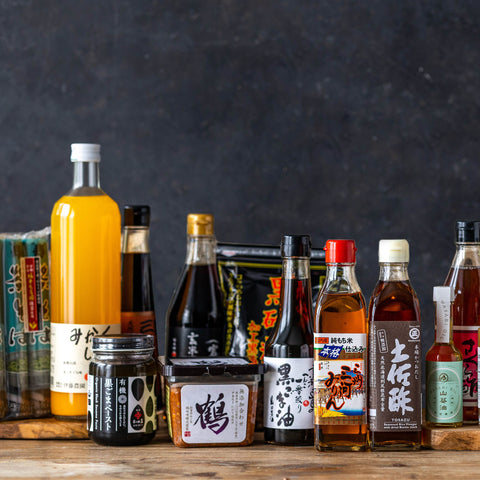 A variety of Japanese condiments, sauces, and packaged foods are arranged on a wooden surface against a dark background.