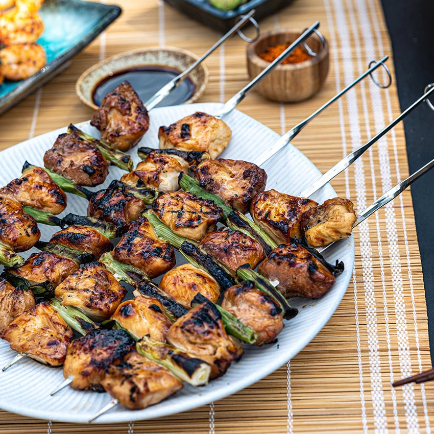 Grilled chicken and vegetable skewers on metal sticks served on a white plate, with sauces and side dishes in the background.