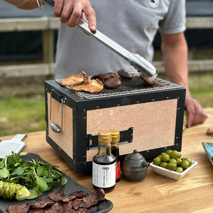A person grills meat and mushrooms on a small tabletop charcoal grill, with assorted sauces, olives, and greens arranged on the wooden table nearby.