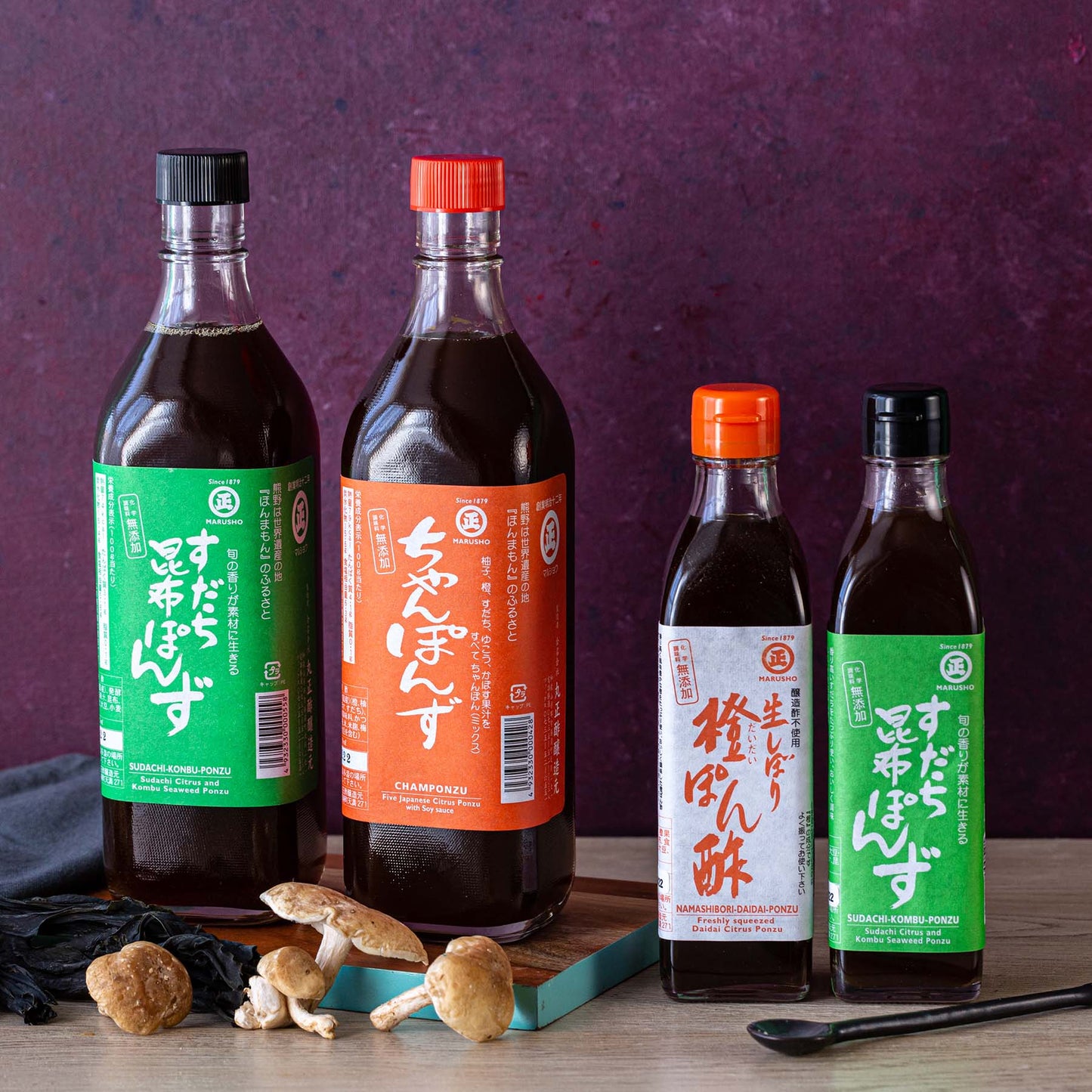 Four bottles of Japanese ponzu sauce are arranged on a tabletop with mushrooms, a black cloth, and a teal chopstick, set against a burgundy background.