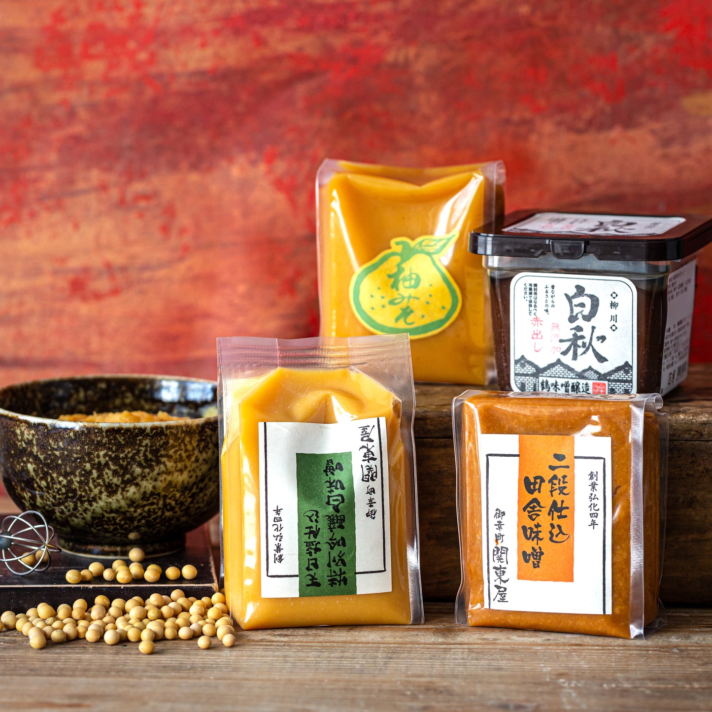 Assorted Japanese miso pastes in plastic packaging and a black container, arranged with soybeans and a rustic bowl against a red and wooden background.