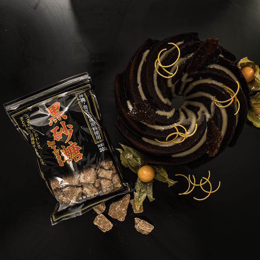 A swirl chocolate cake with white glaze and orange zest, garnished with physalis, beside a bag of Japanese brown sugar pieces on a black surface.
