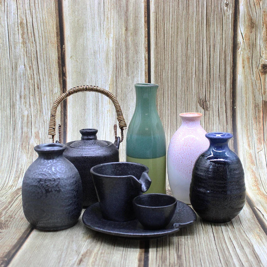 A collection of ceramic vessels, including a teapot, cups, vases, and jars, is arranged on a wooden surface with a wood panel background.