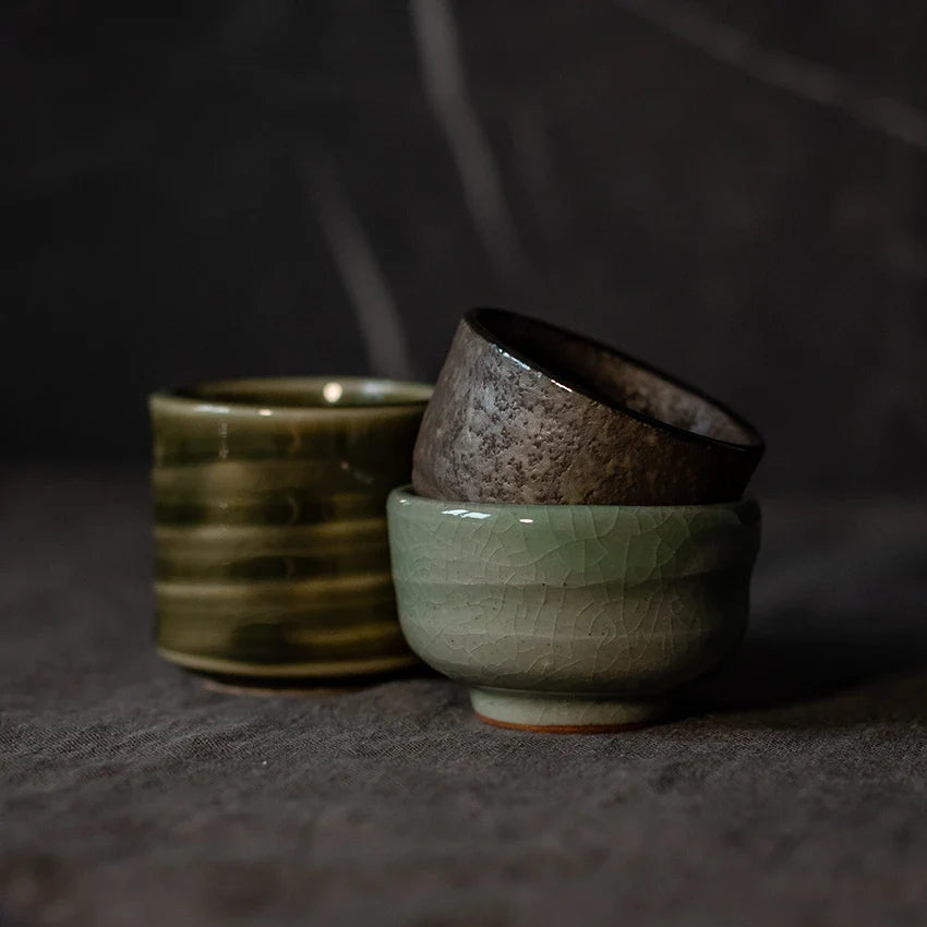 Three ceramic tea cups in green, brown, and gray tones are stacked on a dark surface with a dark background.