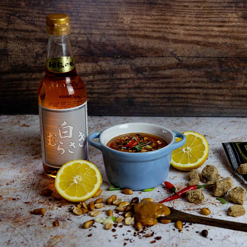 A bottle of mirin, a bowl of sauce with herbs and chili, lemon halves, peanuts, brown sugar, and a wooden spoon with sauce are arranged on a rustic surface.