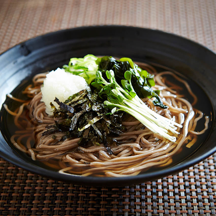 A black bowl filled with soba noodles in broth, topped with grated daikon, seaweed strips, and green garnishes, on a woven placemat.