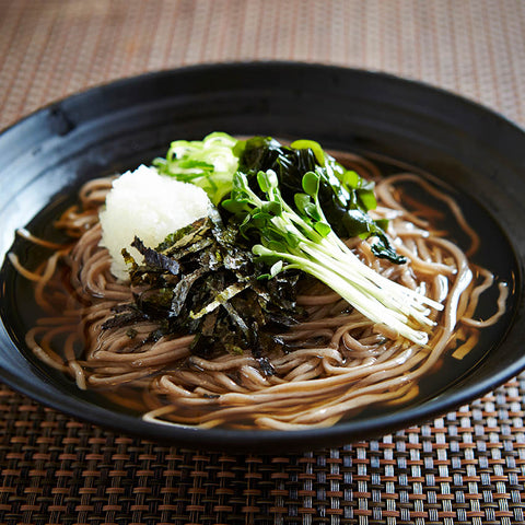 A black bowl filled with soba noodles in broth, topped with grated daikon, seaweed strips, and green garnishes, on a woven placemat.