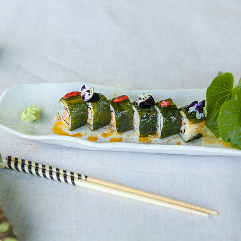 A rectangular plate with five pieces of sushi wrapped in green seaweed, garnished with sauce and edible flowers, served with wasabi and chopsticks on the side.