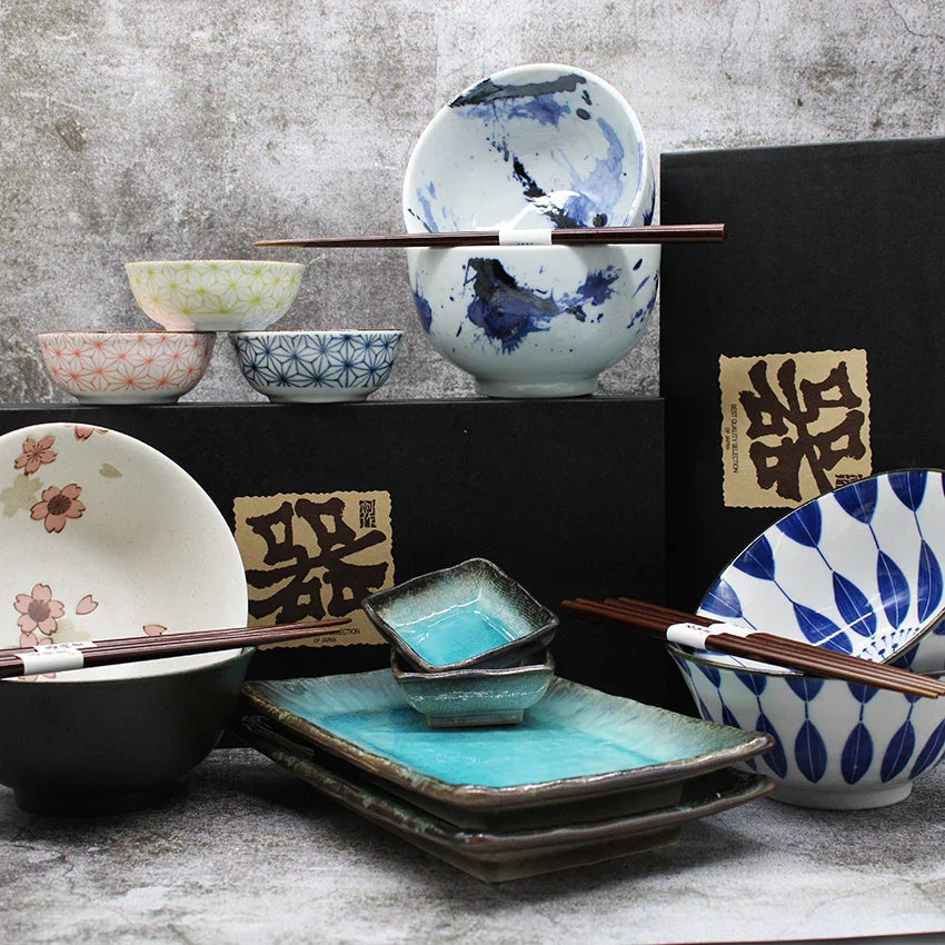 A collection of Japanese ceramic bowls, plates, and dishes with various patterns, two pairs of chopsticks, and decorative black boxes in the background.