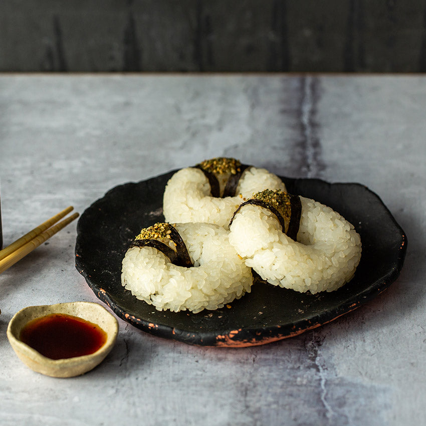 Three sushi pieces with white rice and seaweed are arranged on a black plate, with chopsticks and a small dish of soy sauce beside them.