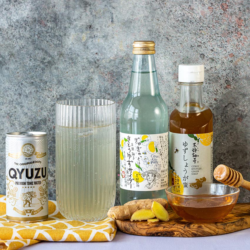A can, a glass, a bottle, and a jar of Yuzu drinks and condiments are arranged with ginger, honey, and a yellow cloth on a stone surface.