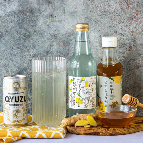A can, a glass, a bottle, and a jar of Yuzu drinks and condiments are arranged with ginger, honey, and a yellow cloth on a stone surface.
