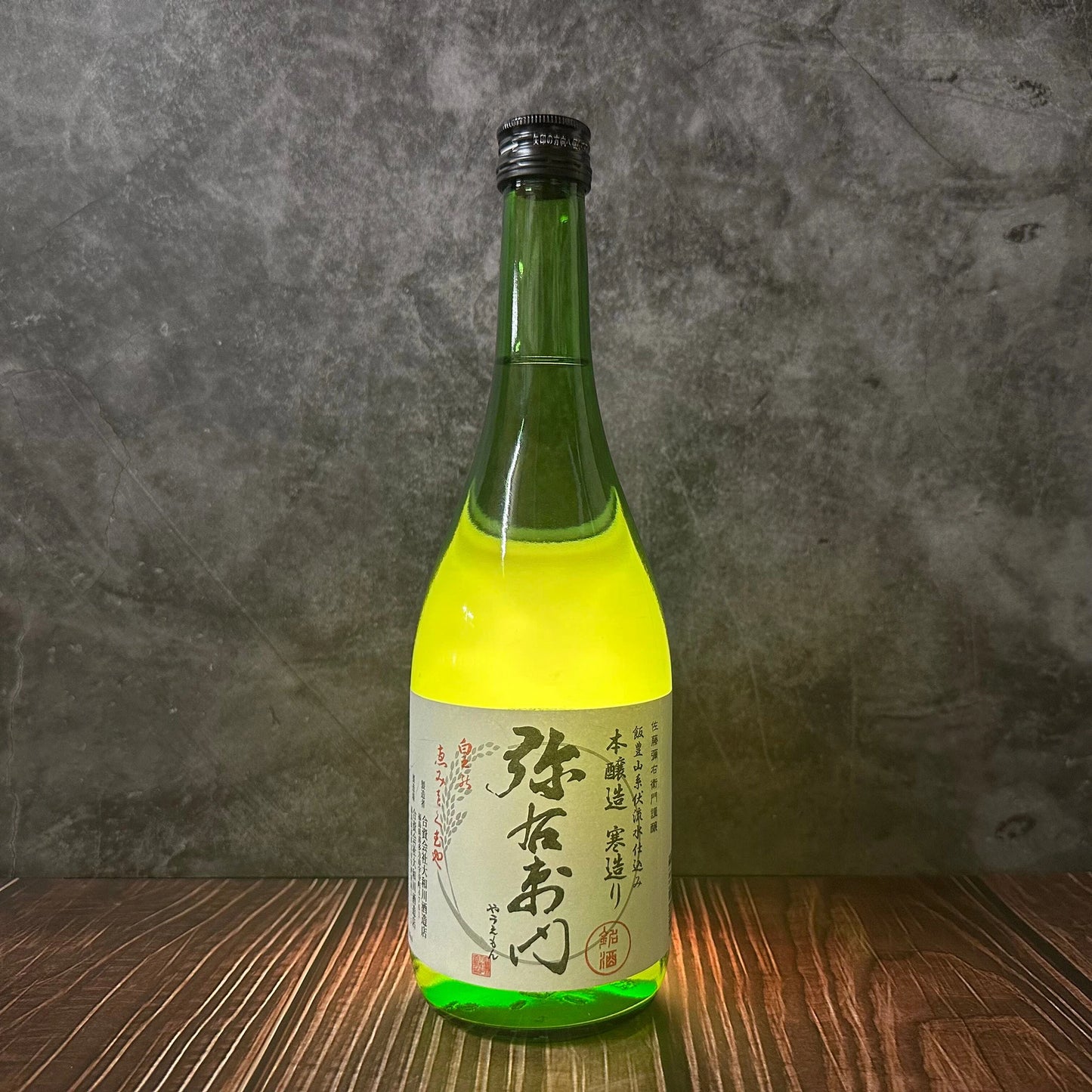 A green glass bottle of Autumn Leaves - Honjozo Sake with a white Japanese label rests on a wooden surface against a gray textured background, evoking the creamy rice notes within.