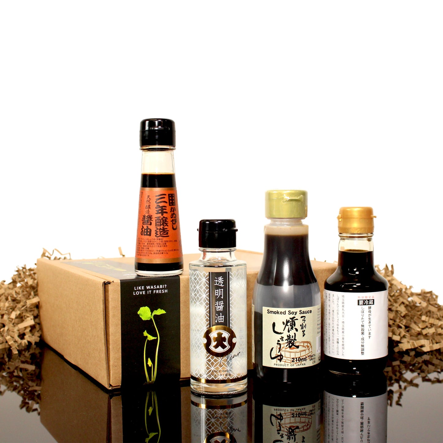Four bottles from the Connoisseurs Soy Sauce Collection, featuring Japanese sauces like Nama Shoyu and Smoked Soy Sauce, are arranged in front of a gift box with decorative paper fill on a reflective surface.