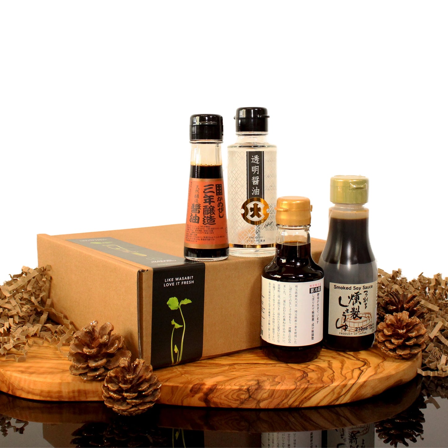 The Connoisseurs Soy Sauce Collection, a set of four assorted Japanese soy sauces including Nama Shoyu and Smoked Soy Sauce, is displayed on a wooden board with pinecones beside a brown gift box labeled wasabi against a white background.