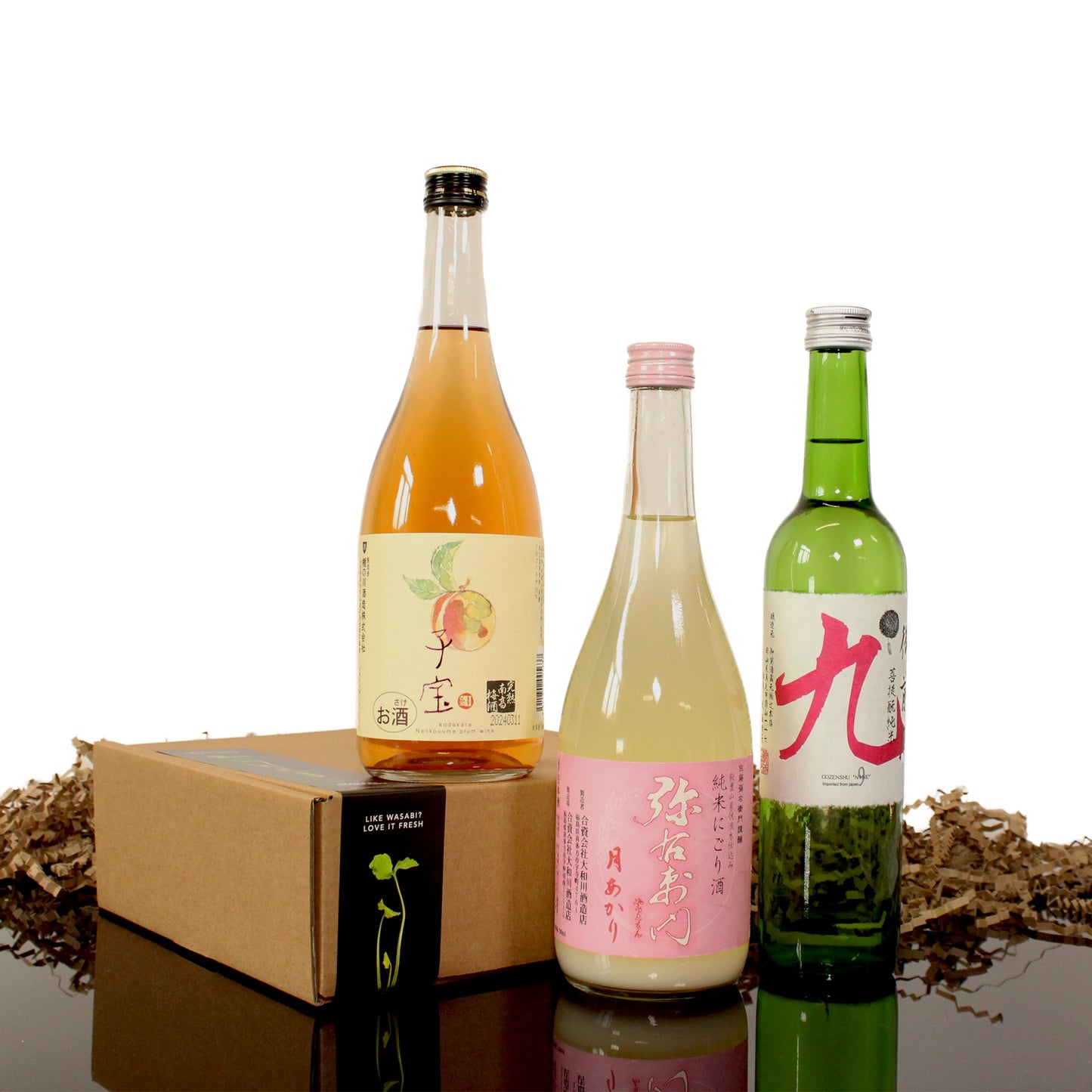 The Deluxe Sake Selection features three bottled Japanese sake and umeshu with unique labels, displayed next to a closed brown gift box filled with shredded paper on a clean white background.
