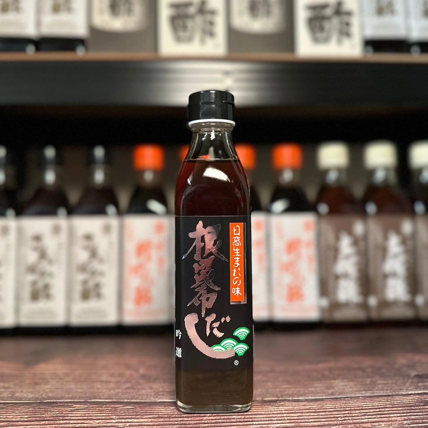 A bottle of Extra Ne Kombu Concentrated Dashi (300ml) with a black label and white and red text sits on a wooden surface, evoking rich umami flavors, while similar bottles appear blurred in the background.