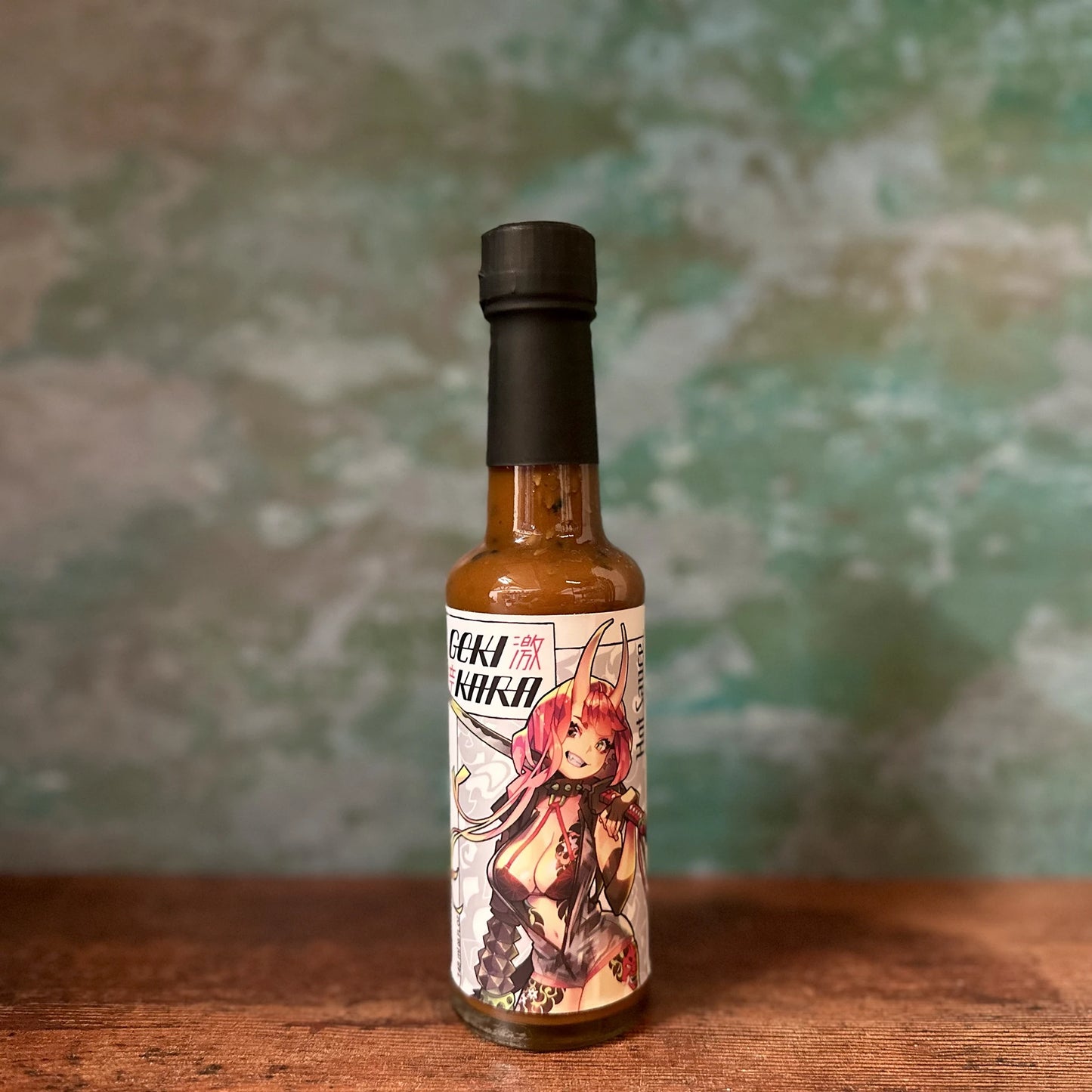 A 148ml bottle of Gekikara Hot Sauce with an anime-style character on the label is placed on a wooden surface, set against a mottled green and gray background.