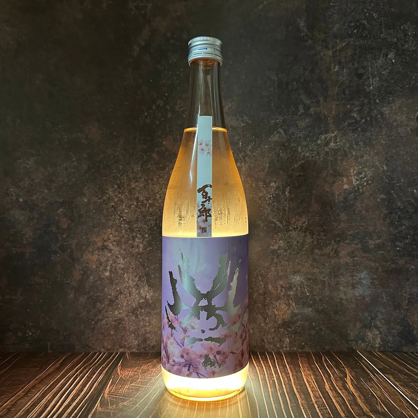 Hyakujuro Sakura Junmai Daiginjo (720ml), in a clear glass bottle with a floral sakura label and silver cap, contains light golden sake and stands on a wooden surface with a dark textured backdrop.