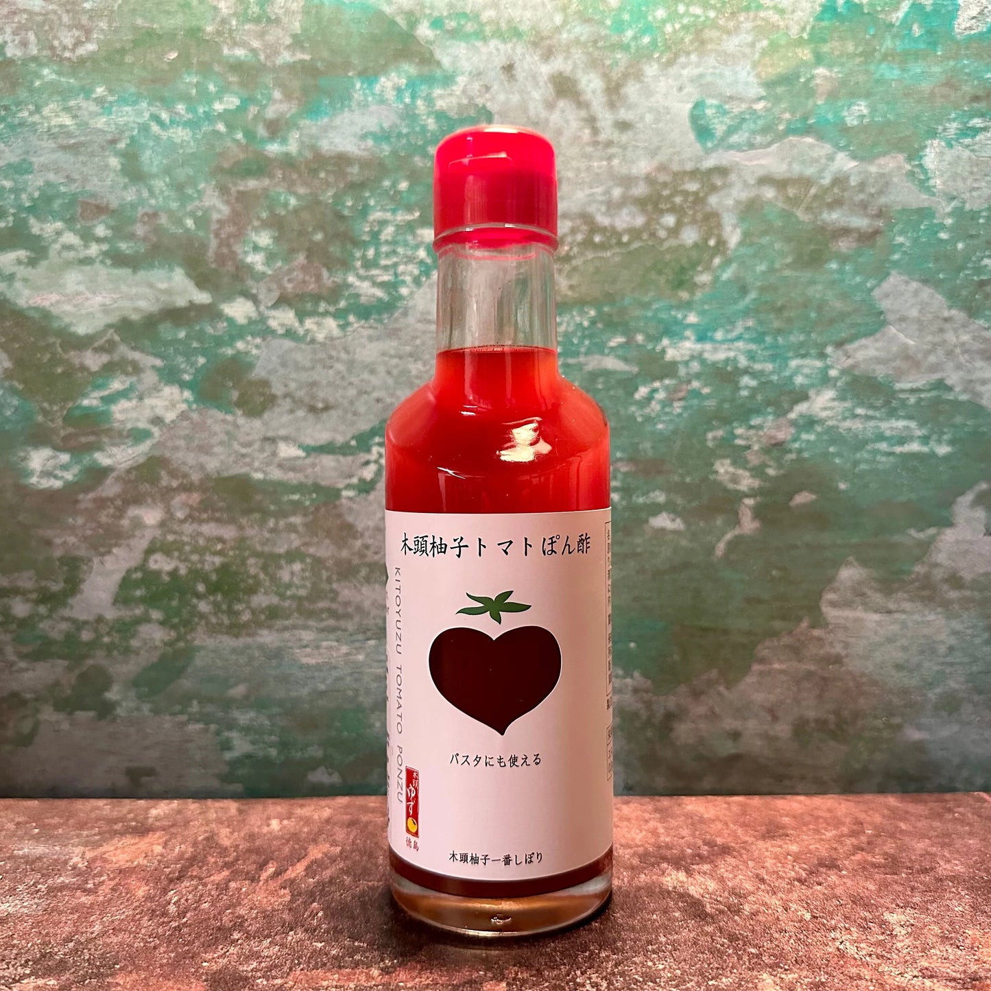 A 200ml glass bottle of Kito Yuzu & Tomato Ponzu, featuring a label with a large tomato illustration and Japanese text, evokes the charm of classic Japanese condiments against a textured backdrop.