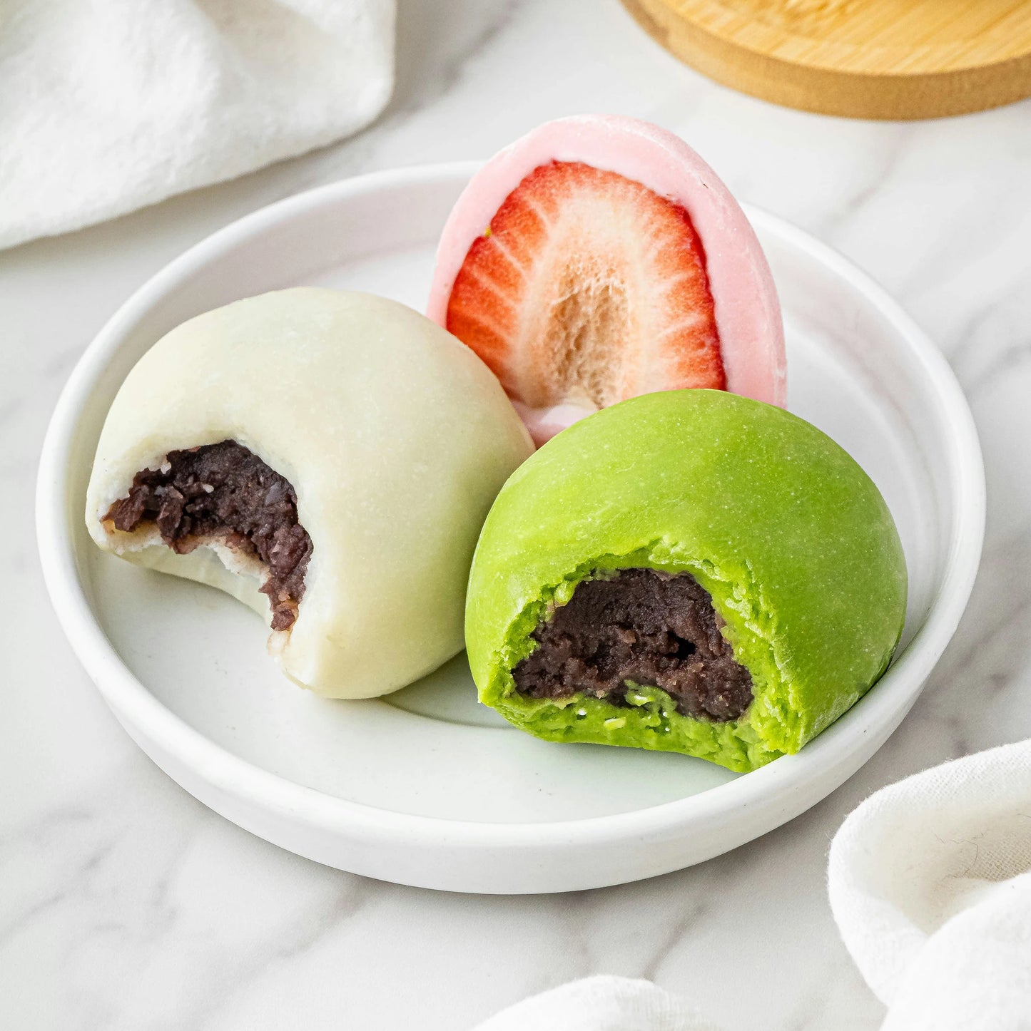Three Japanese desserts on a white plate: pink strawberry mochi, plus white and green varieties made with Mochiko Komeko Rice Flour for Mochi (300g), both filled with red bean paste and showing bite marks.
