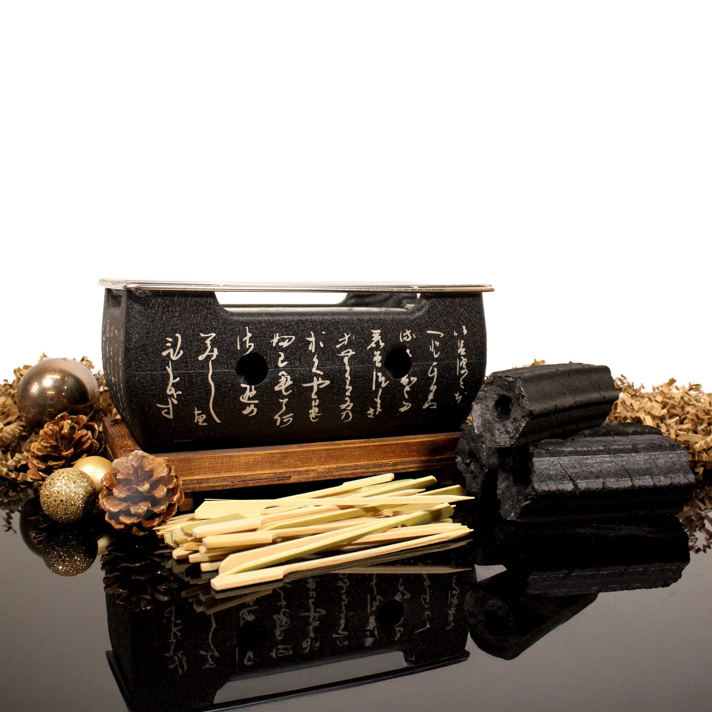 The Konro Aluminium Grill 24cm Barbecue Set features bamboo skewers, Binchotan charcoal pieces, a pine cone, and decorative balls, all arranged with white script on a reflective surface.