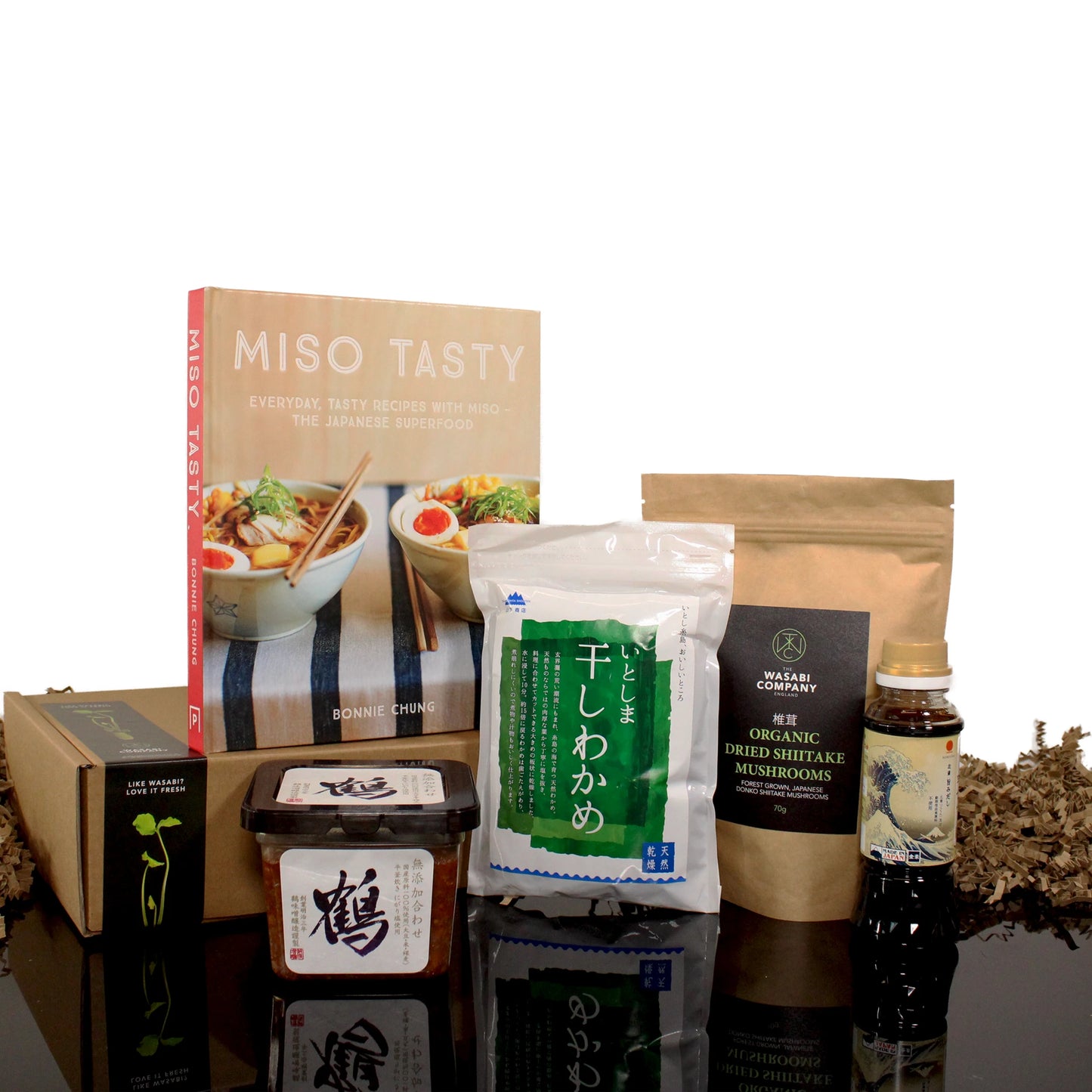 The Miso Soup Kit with Miso Tasty Cook Book includes a cookbook, dried wakame, dried shiitake mushrooms, Japanese miso paste, soy sauce, and seasoning—all beautifully displayed on a black surface.