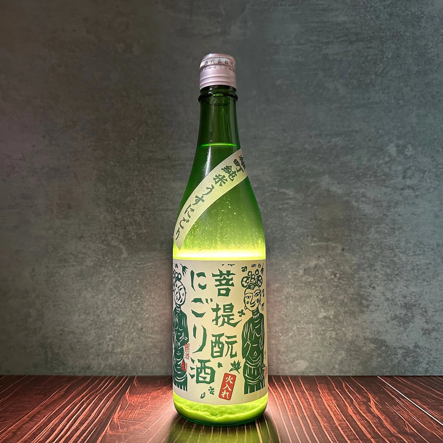 A 720ml bottle of Misty Mountain Junmai Bodaimoto Nigori Sake with a white and green label featuring Japanese text and illustrations sits on a wooden surface against a gray background.
