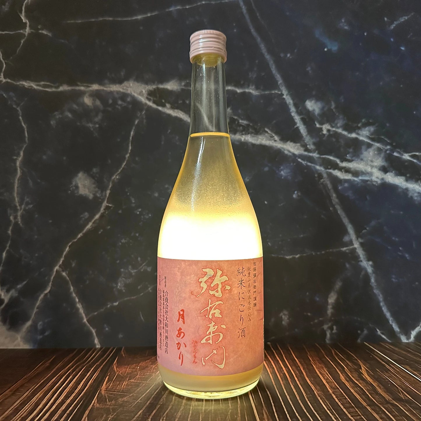 A clear glass bottle of Moonlight Junmai Nigori sake (720ml) with a light yellow hue and a pink Japanese label sits on a dark wood surface against a marbled dark background.