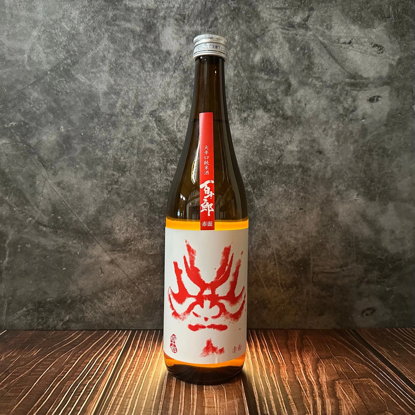 A brown glass bottle of Hyakujuro Red Face - Junmai (Karakuchi) Sake with a red and white crab label stands on wood against a gray textured background, suggesting its versatile sake and subtle rice flavors.