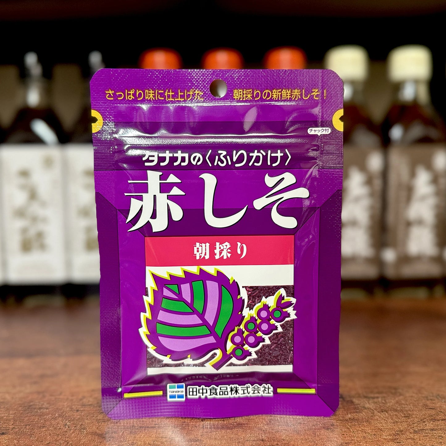 A purple package of Red Shiso Furikake Tanaka (22g), a Japanese seasoning made from red shiso leaves, is shown on a wooden surface with blurred bottles in the background.