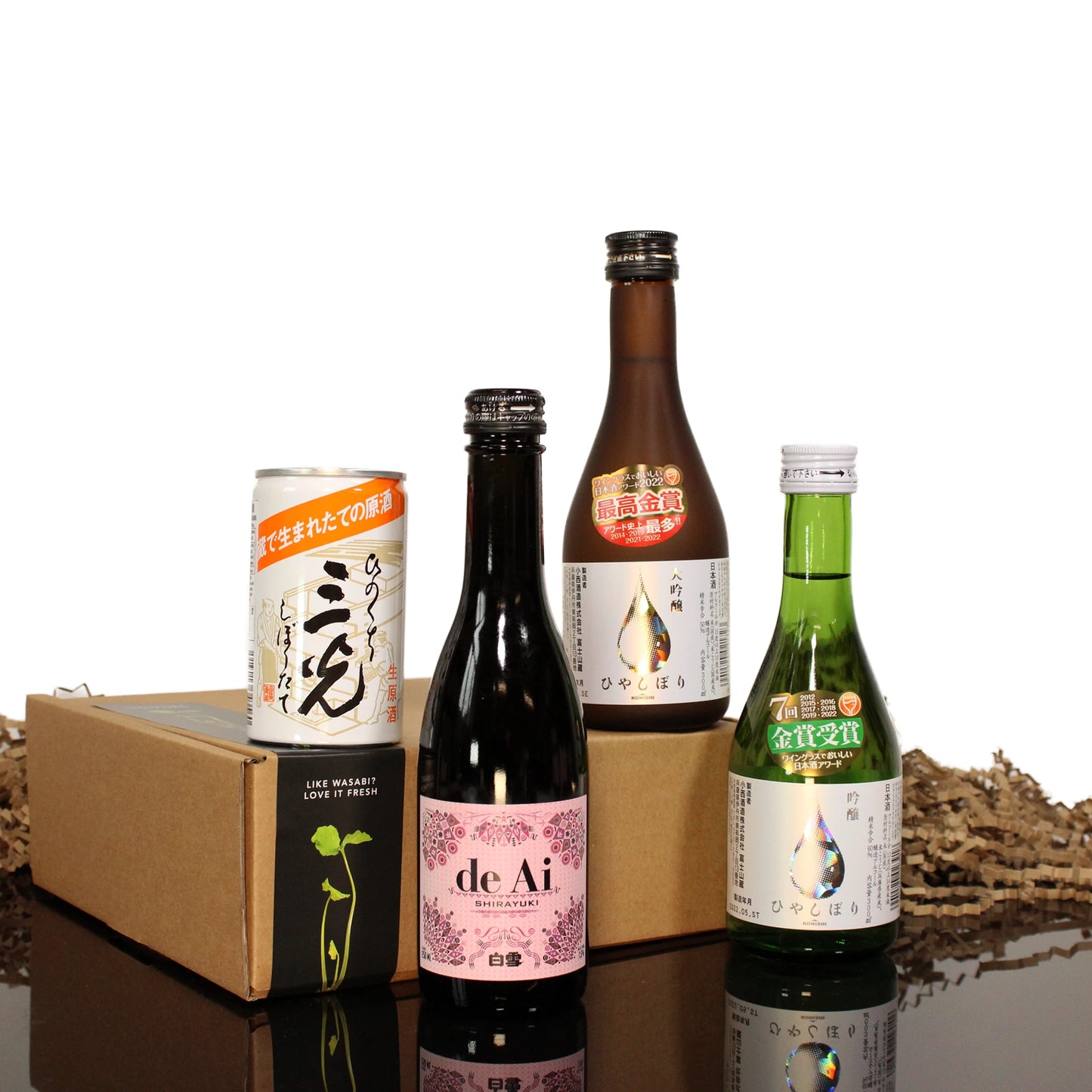Four bottles and cans from the Sake Selection are arranged before a cardboard box, with wood shavings scattered at the base.