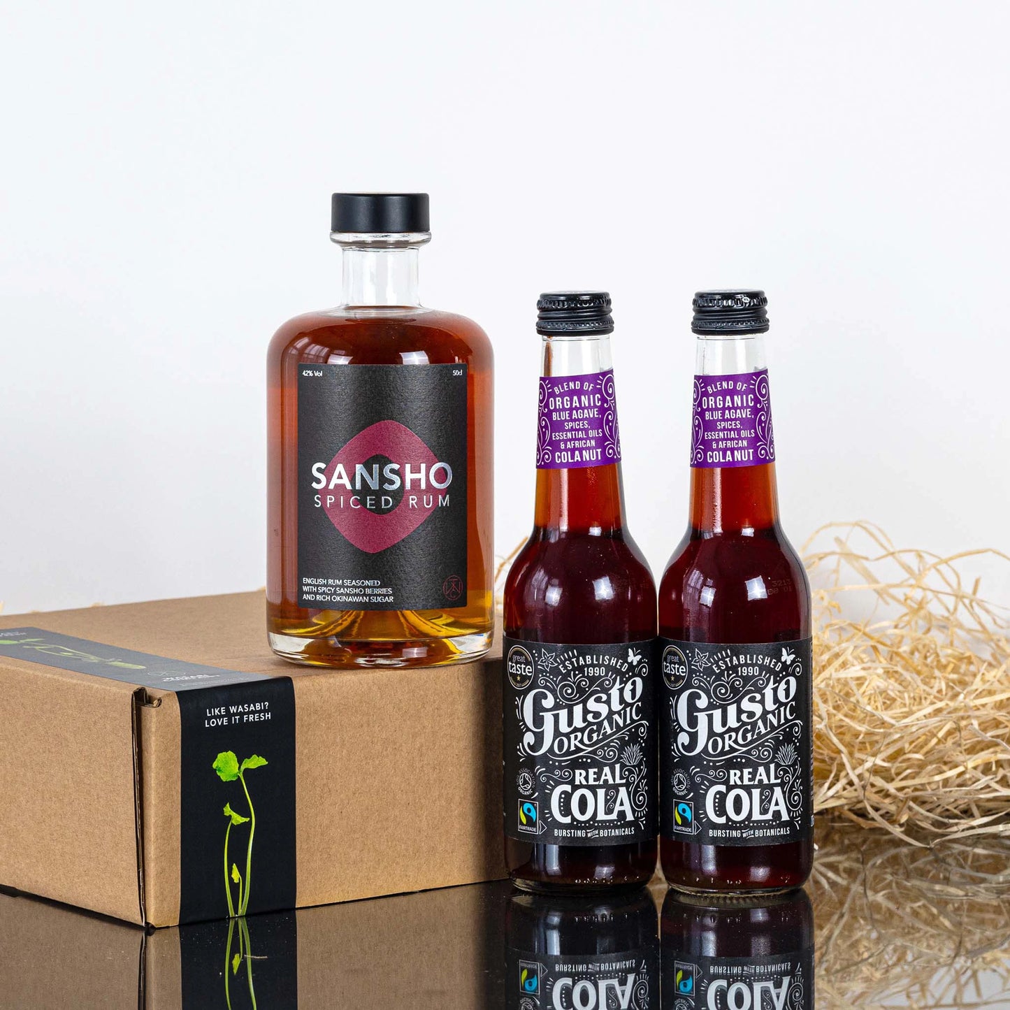 The Sansho Rum & Gusto Cola Set, featuring a bottle of Sansho Spiced Rum and two Gusto Organic Colas, comes neatly packed in a cardboard box with straw on a white surface.