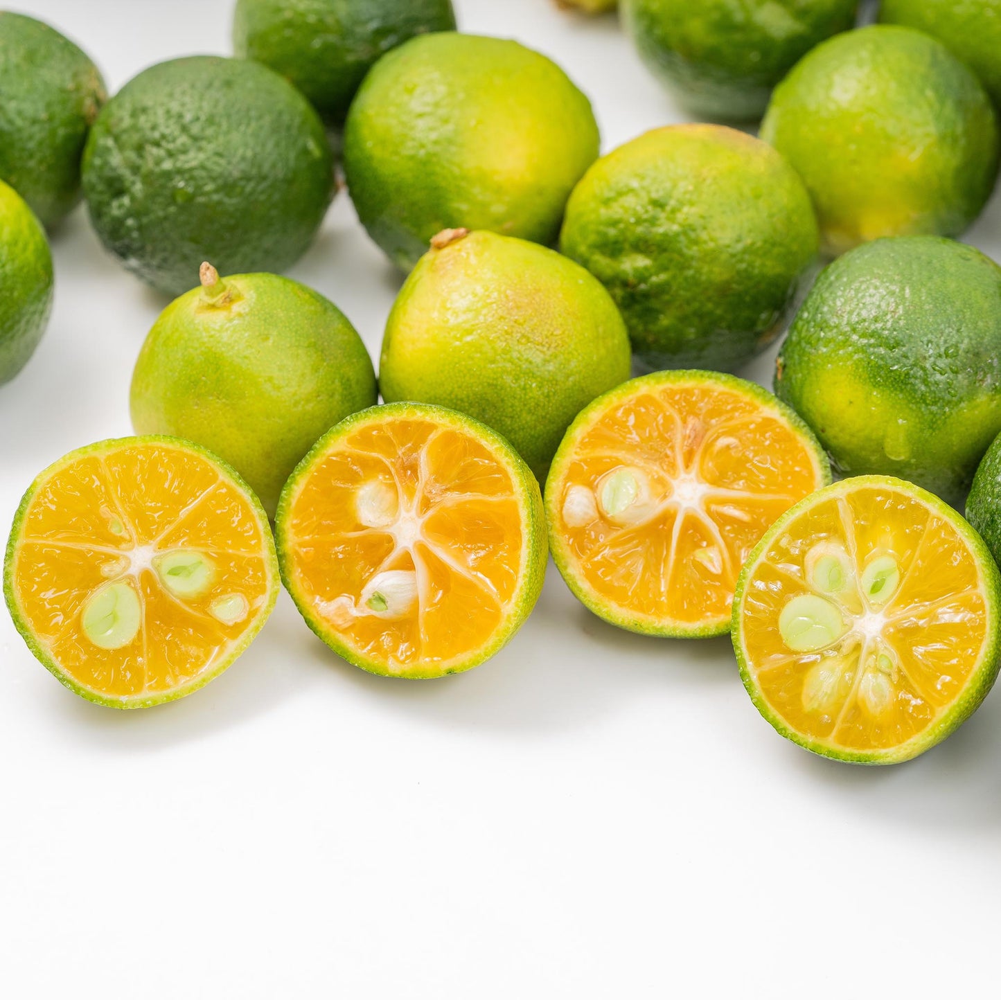 Fresh Shikuwasa Fruit, a zesty citrus popular in Japanese cuisine, is shown whole and halved on a white surface, revealing green rinds and bright orange interiors with seeds.