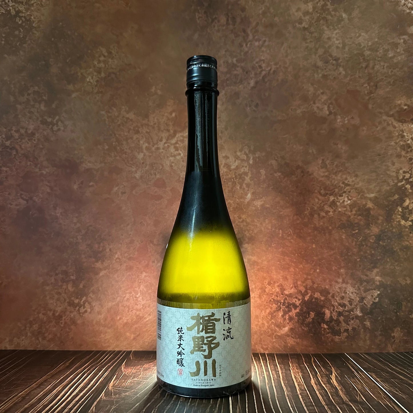 A single bottle of Stream Junmai Daiginjo Sake - 720ml stands on a dark wooden surface, featuring a beige label with Japanese writing and set against a brown textured background.
