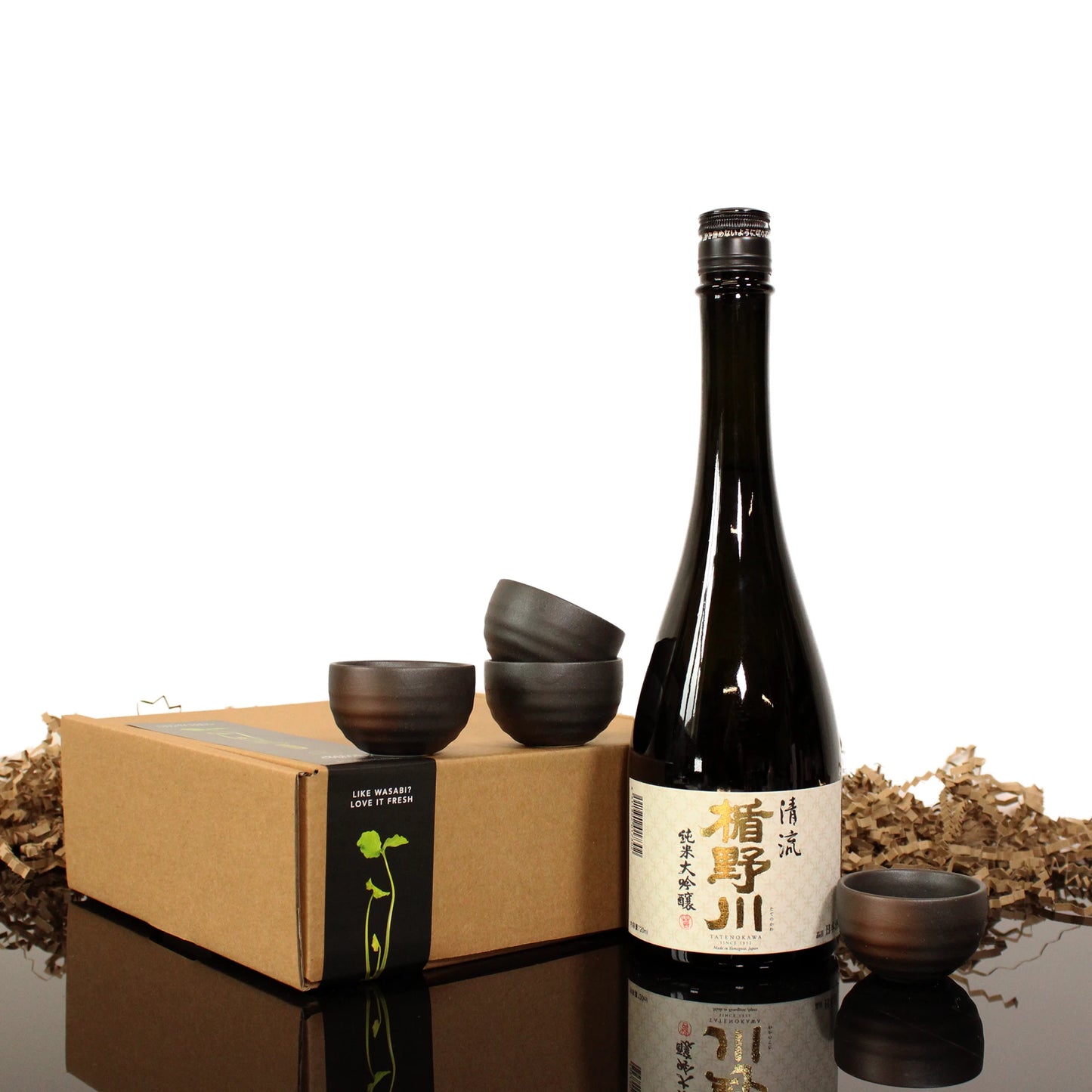 A Stream Sake black Junmai Daiginjo bottle with Japanese script, four elegant sake cups, and a brown gift box sit on a reflective surface with packing material.
