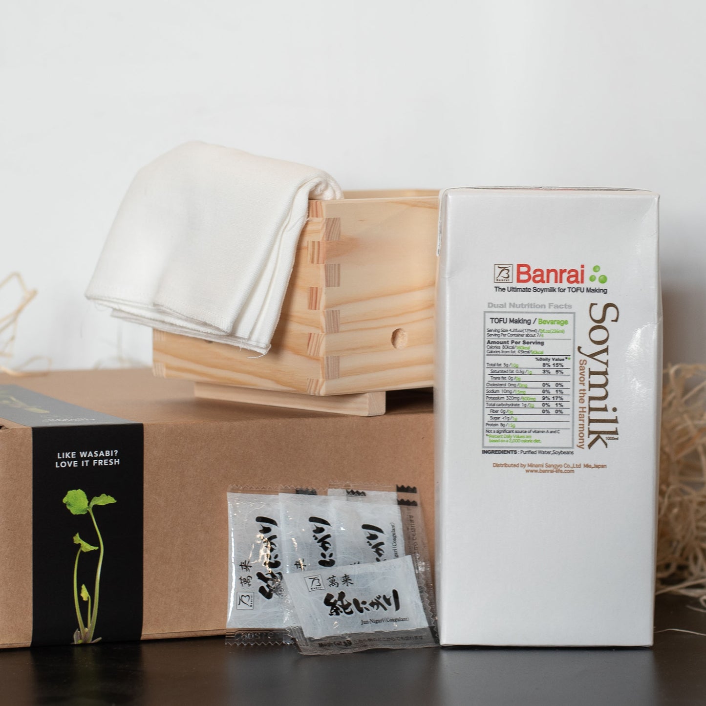 The Tofu Making Kit, featuring Banrai soymilk, a tofu press with cloth, condiment packets, and a black box with wasabi plant art, is neatly arranged—ideal for vegan diets.