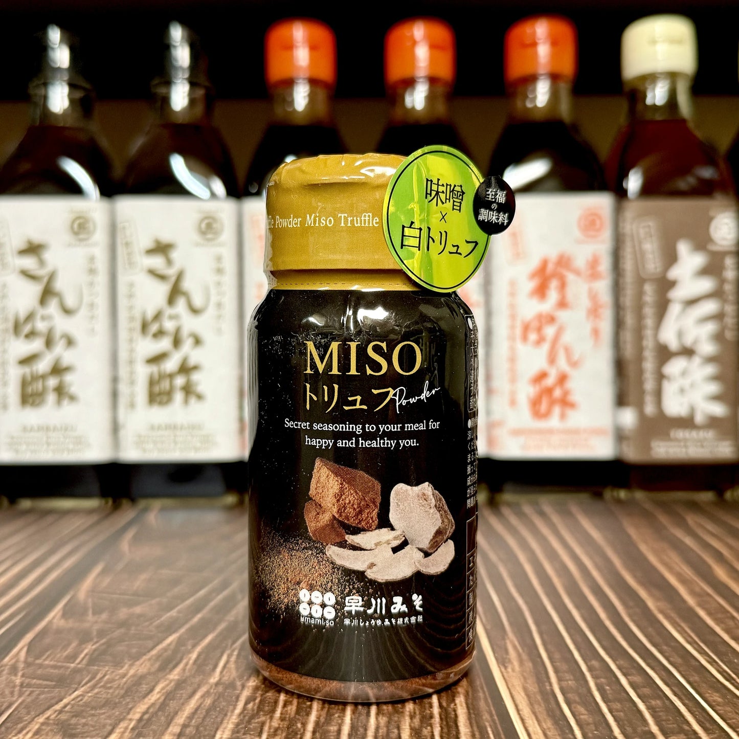 A 70g jar of Umami-so Miso Seasoning with Truffle, made with bianchetto truffles, rests on a wooden surface, while bottles of assorted Japanese sauces are arranged in the background.