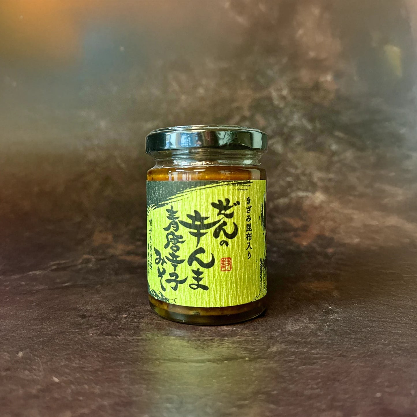 A 120g jar of Vegan Zen no Karanma Condiment with a green Japanese label sits on a dark surface and blurred brown background—this vegan Japanese condiment delivers rich umami flavor, ideal for enhancing your favorite dishes.