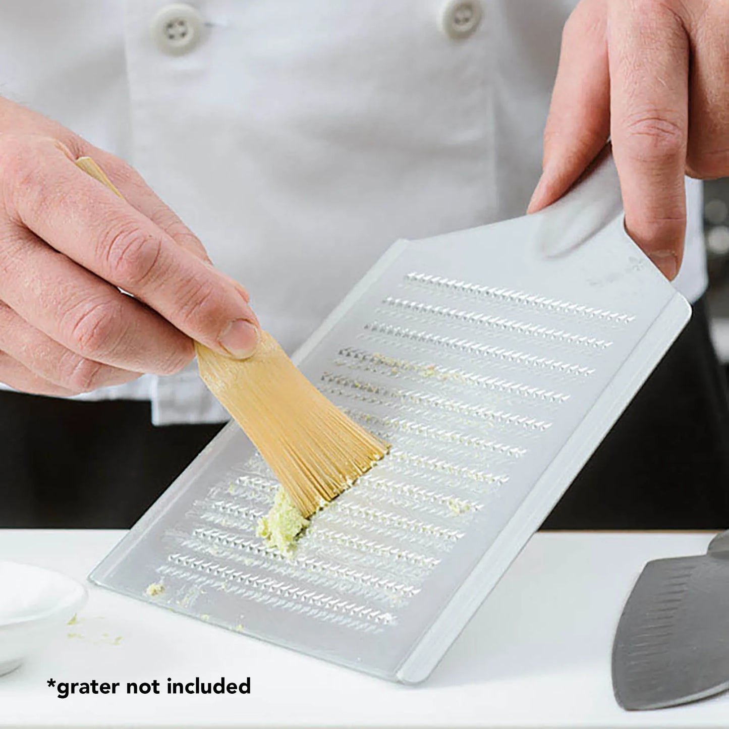 A person uses a Bamboo Wasabi Grater Brush to collect grated food from a flat metal grater held over a counter. Text in the image reads *grater not included.