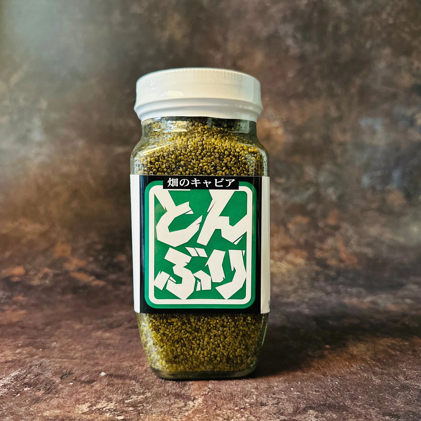A clear plastic jar with a white lid contains green Tonburi granules (vegan caviar). The label reads Watanabe Tonburi - 280g in Japanese, white text on green. The jar is placed on a brown textured surface.