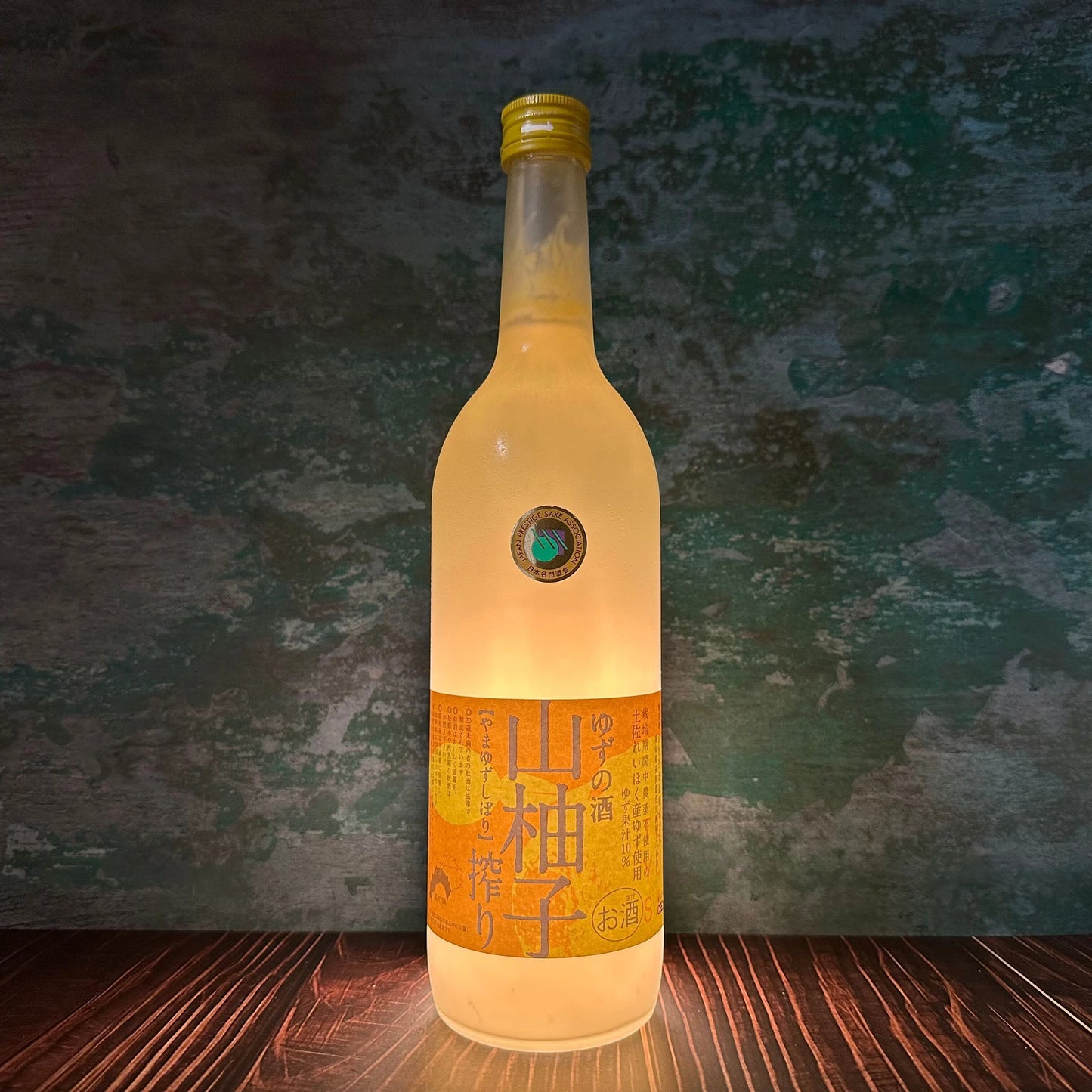 A 720ml bottle of Yama Yuzu Shibori Sake, featuring a yellow label, stands upright on a wooden surface with backlighting against a textured green background.
