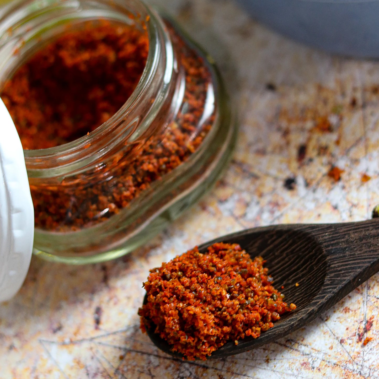 A glass jar of Yuzu Kararin Yuzu Zest & Chilli Mix (35g) lies on its side, spilling some of the vibrant blend onto a wooden spoon placed on a speckled surface.