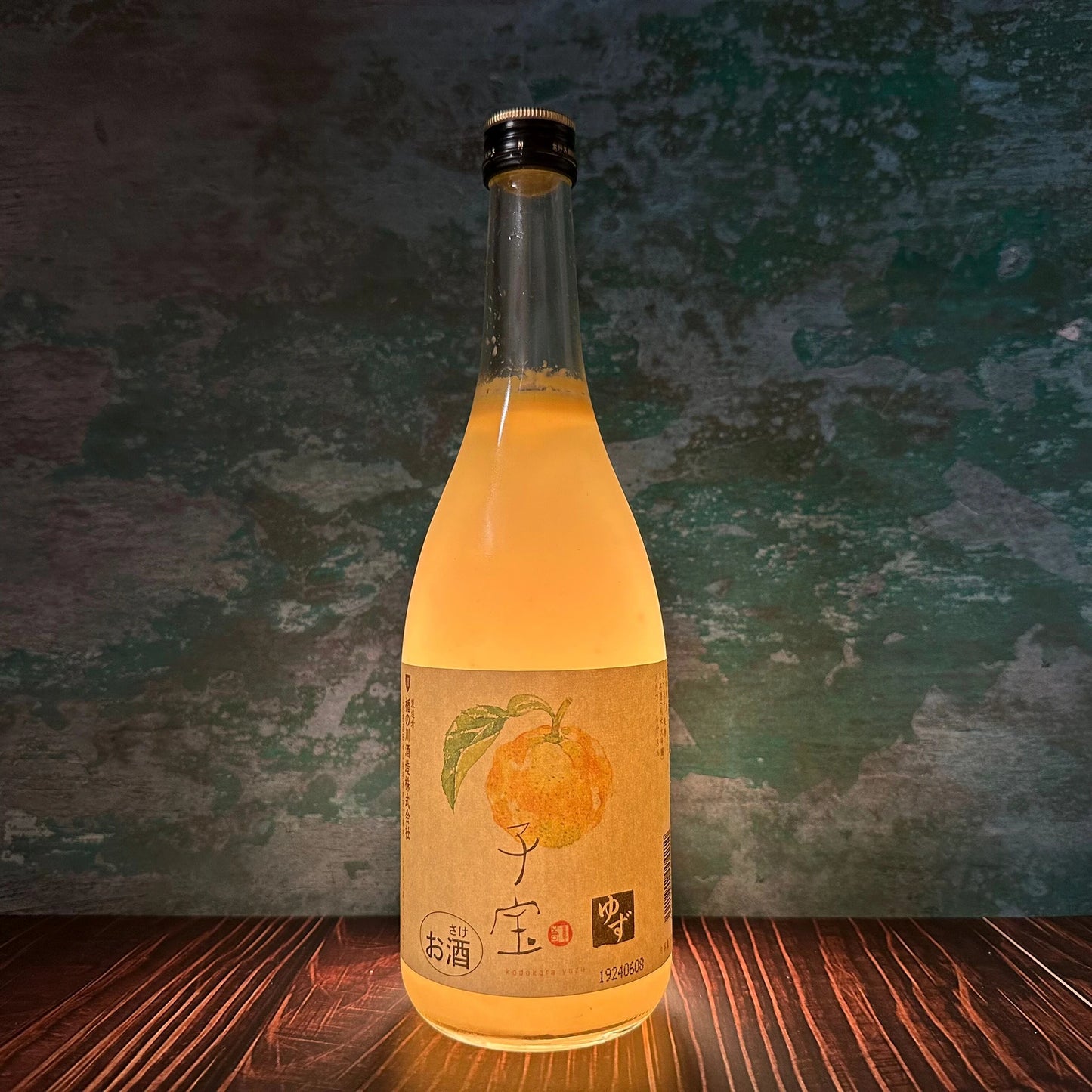 A 720ml bottle of Yuzushu - Yuzu with Shochu, featuring a fruit illustration on the label, sits on a wooden surface against a green and brown textured background.